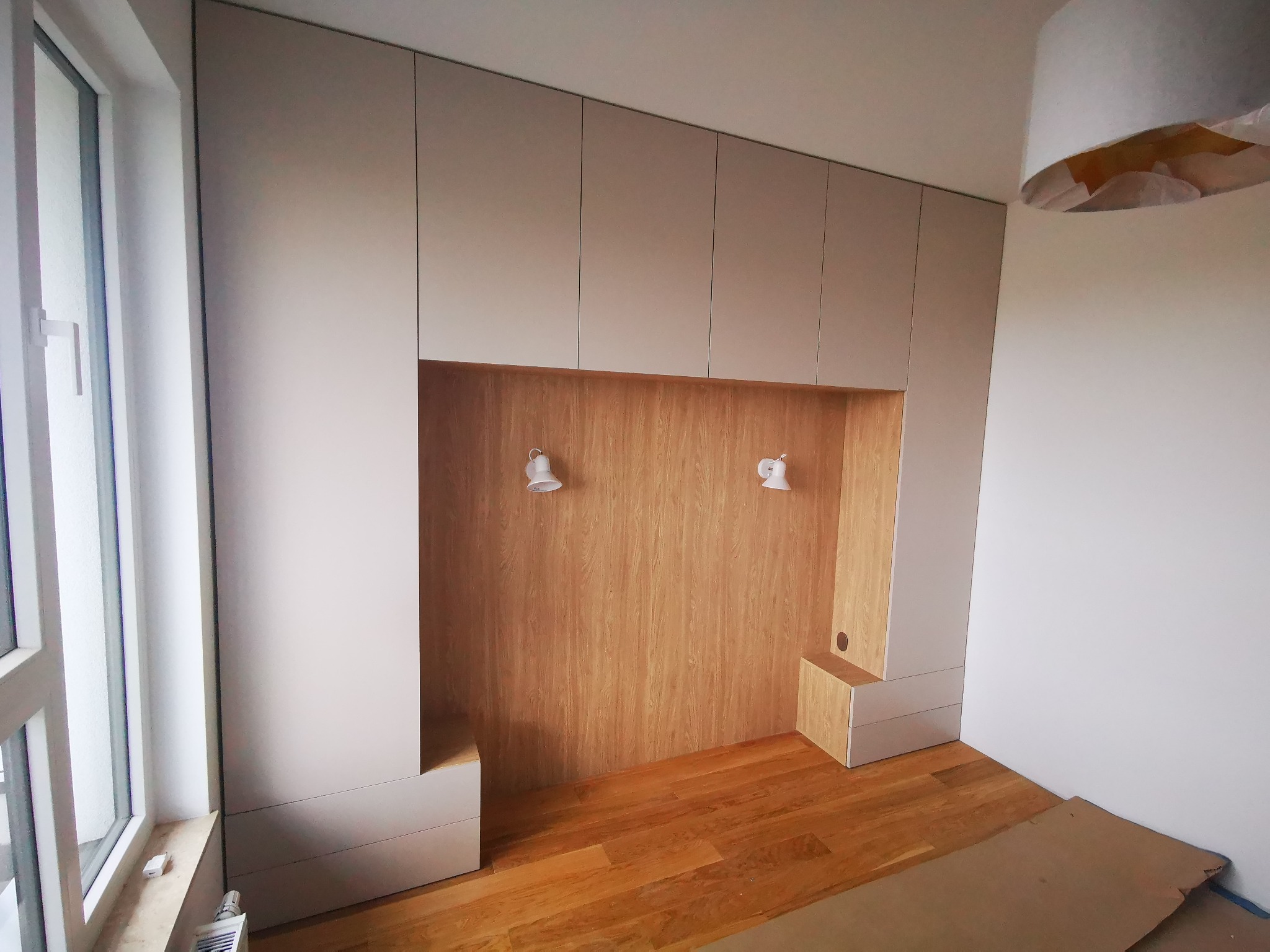 Biało-beżowa zabudowa meblowa z wnęką z drewnianym fornirem, oświetlona dwoma kinkietami, z drewnianą podłogą i dużym oknem po lewej stronie.