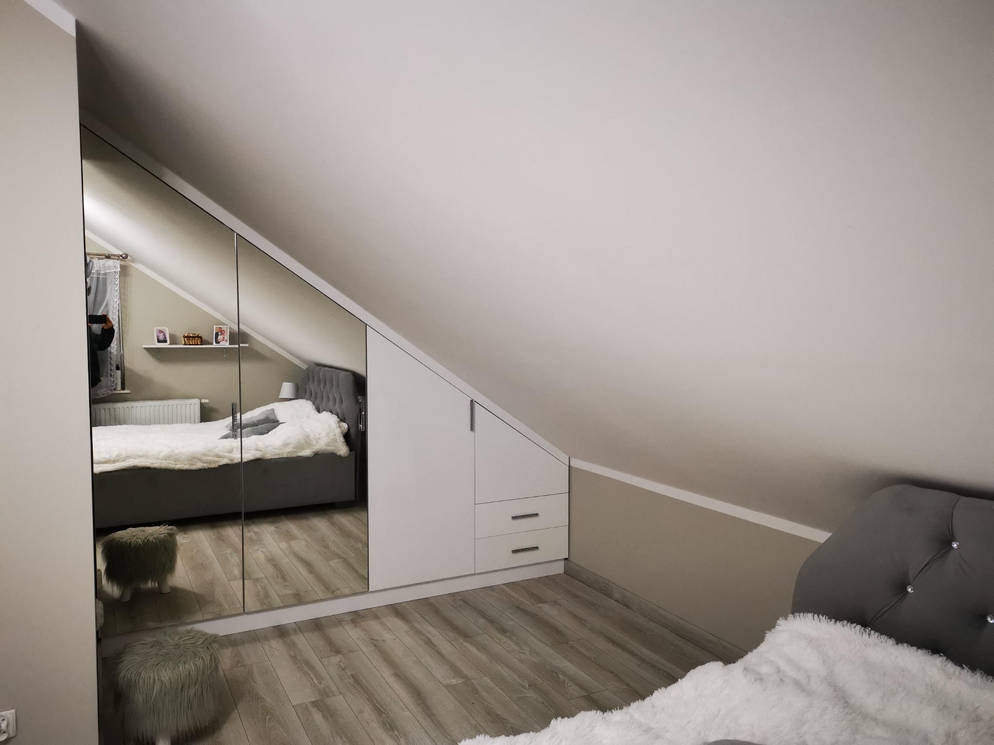 Szafa wnękowa z lustrzanymi drzwiami w pokoju na poddaszu, białe fronty, dwie szuflady, odbicie łóżka z szarym zagłówkiem.