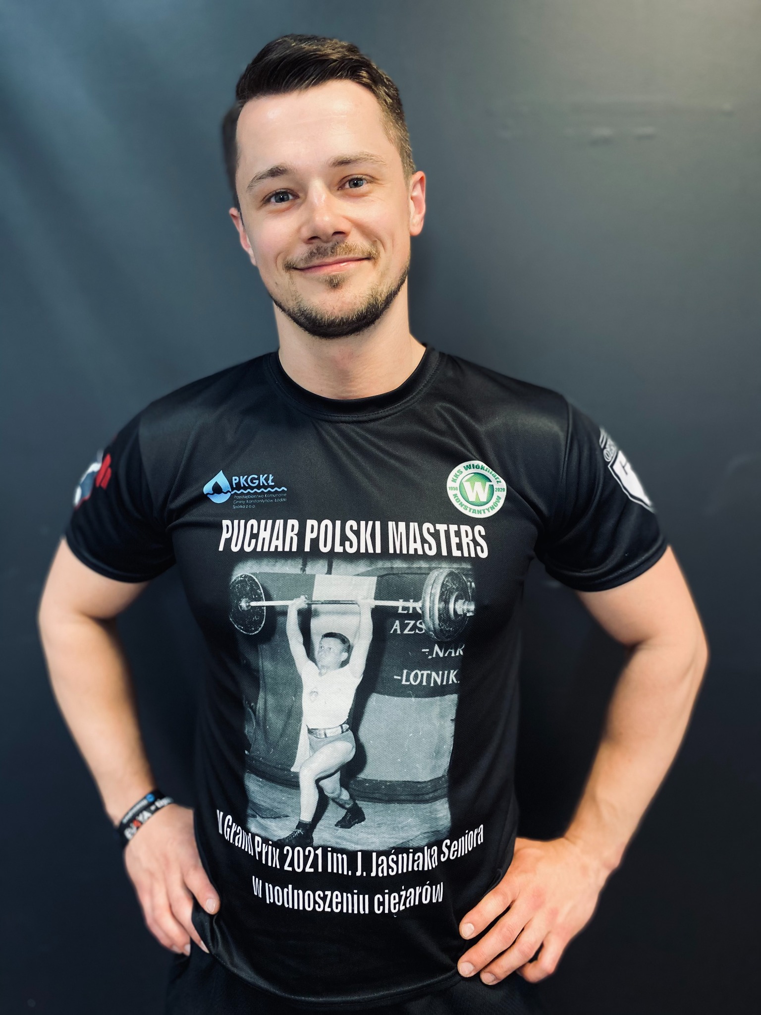 Uśmiechnięty mężczyzna w czarnej koszulce z nadrukiem 'Puchar Polski Masters' i wizerunkiem sztangisty, stojący na tle ciemnej ściany.