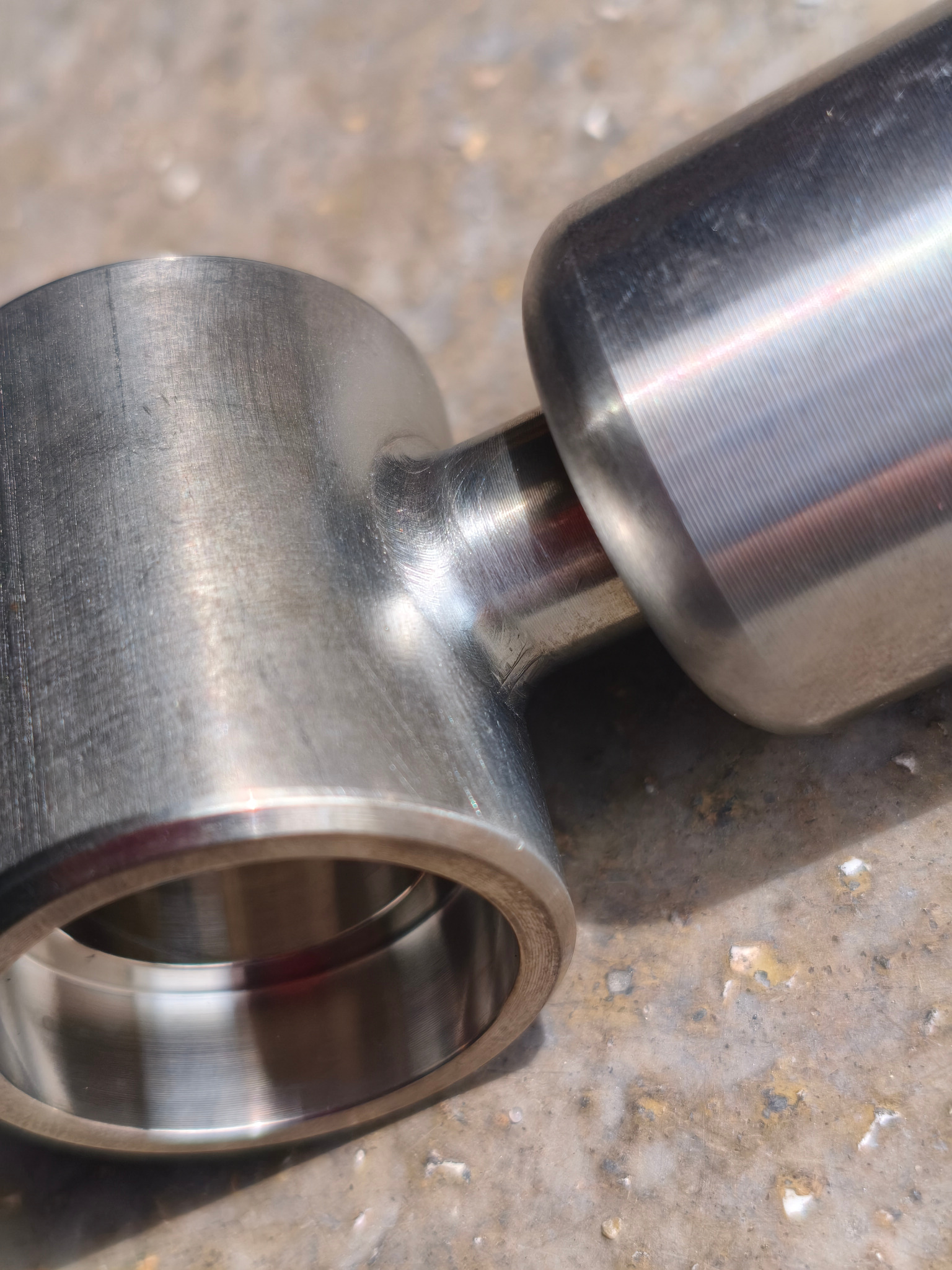 Precyzyjnie spawany, metalowy łącznik rurowy z widocznymi śladami obróbki skrawaniem, leżący na szorstkiej, betonowej powierzchni w silnym oświetleniu.