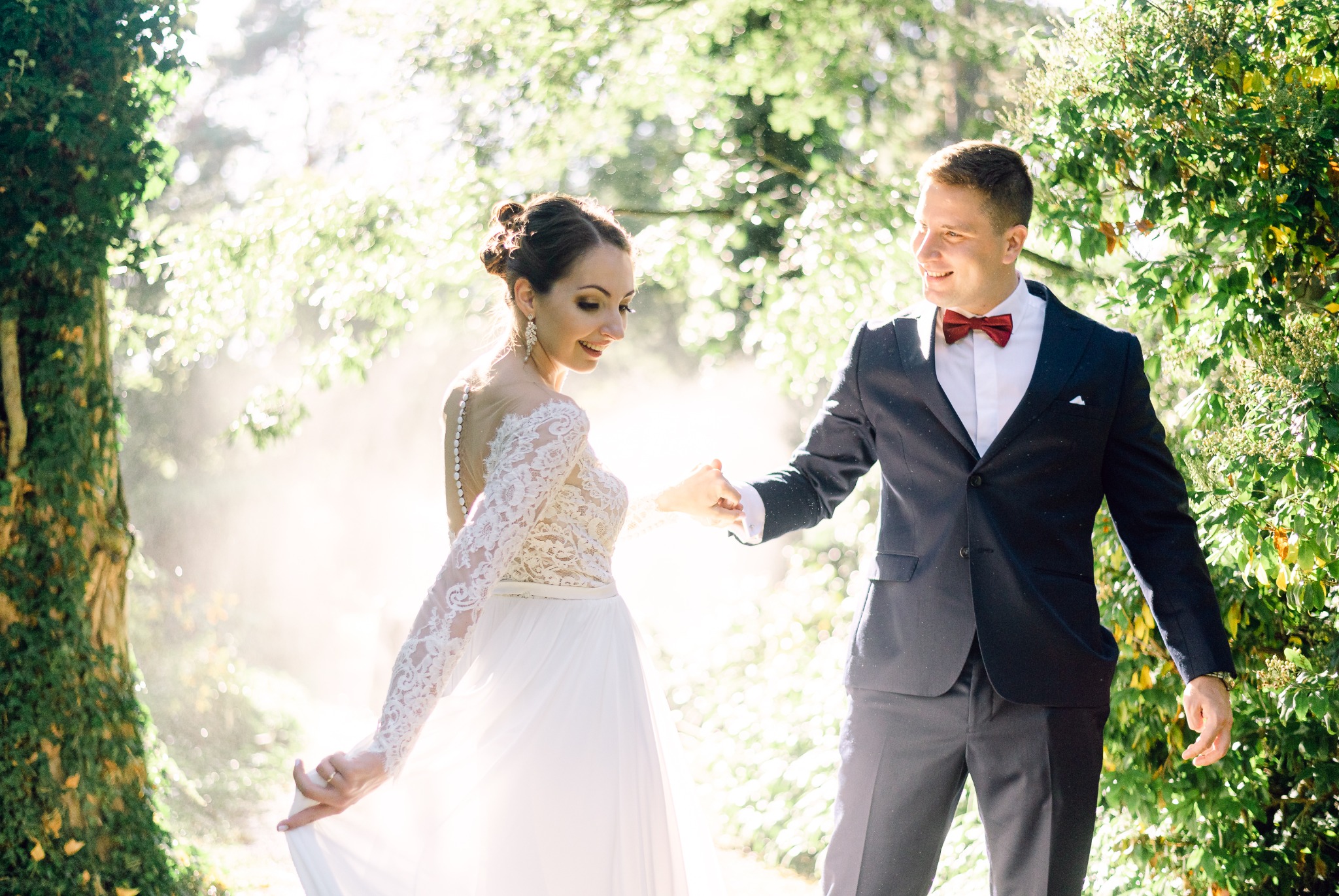 Uśmiechnięta panna młoda w koronkowej sukni i pan młody w granatowym garniturze z bordową muchą trzymają się za ręce w słonecznym lesie, z delikatnym efektem bokeh w tle.