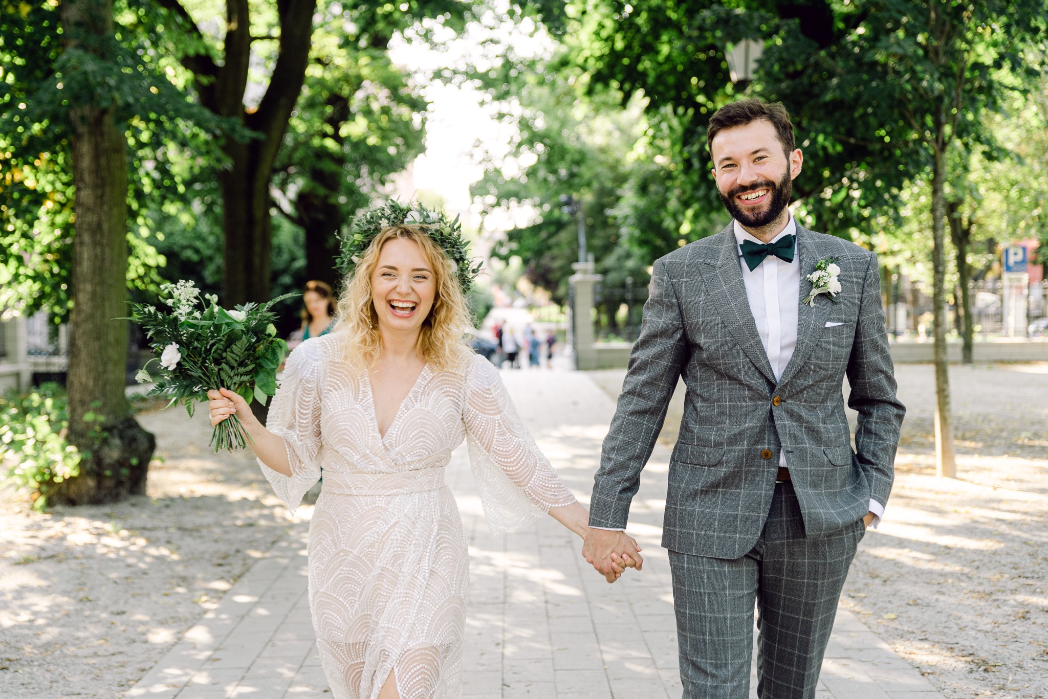 Uśmiechnięta para młoda, on w szarym garniturze w kratę z zieloną muchą, ona w koronkowej sukni z wiankiem, idąca po alei parku trzymając się za ręce.
