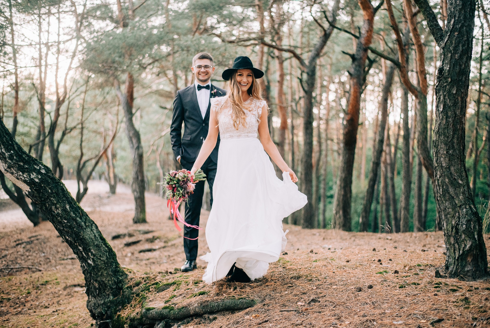 Uśmiechnięta panna młoda w kapeluszu i zwiewnej sukni wiruje na tle leśnej scenerii, za nią stoi elegancki pan młody z bukietem w ręku.