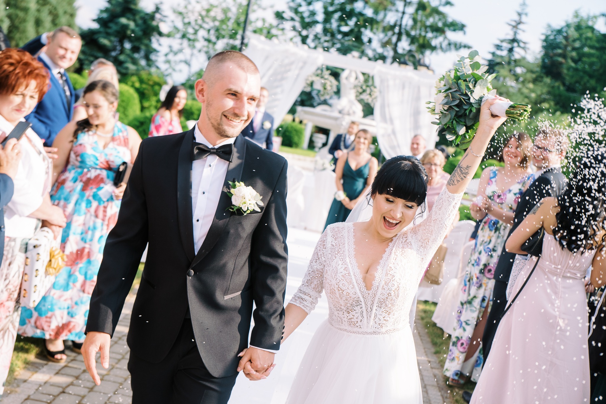 Uśmiechnięta para młoda w trakcie ceremonii zaślubin, idąca trzymając się za ręce, obsypywana konfetti, panna młoda w koronkowej sukni z bukietem uniesionym do góry, pan młody w czarnym smokingu...