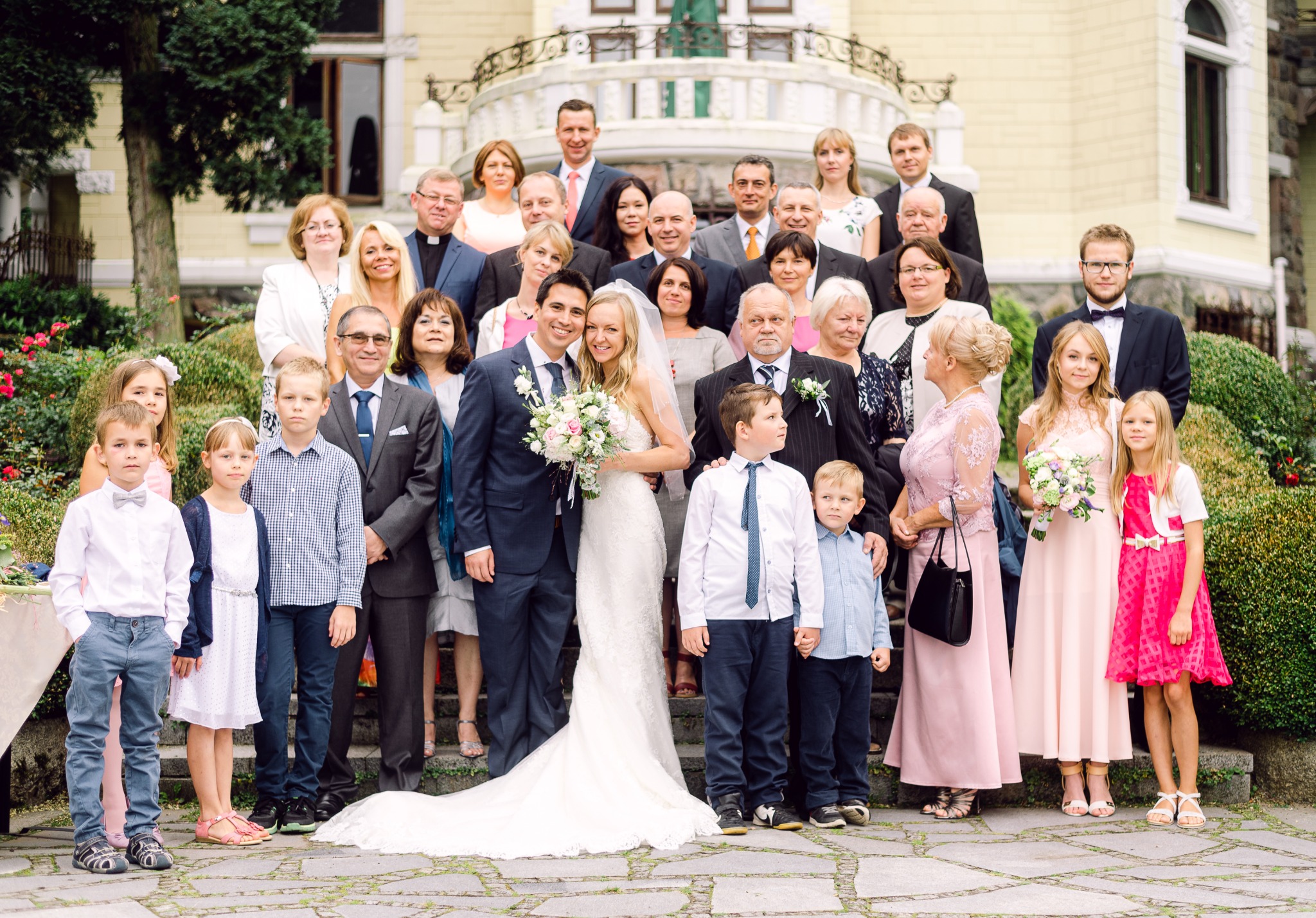 Grupowe zdjęcie ślubne na schodach z panną młodą w koronkowej sukni i panem młodym w granatowym garniturze, otoczonych rodziną i przyjaciółmi w eleganckich strojach.