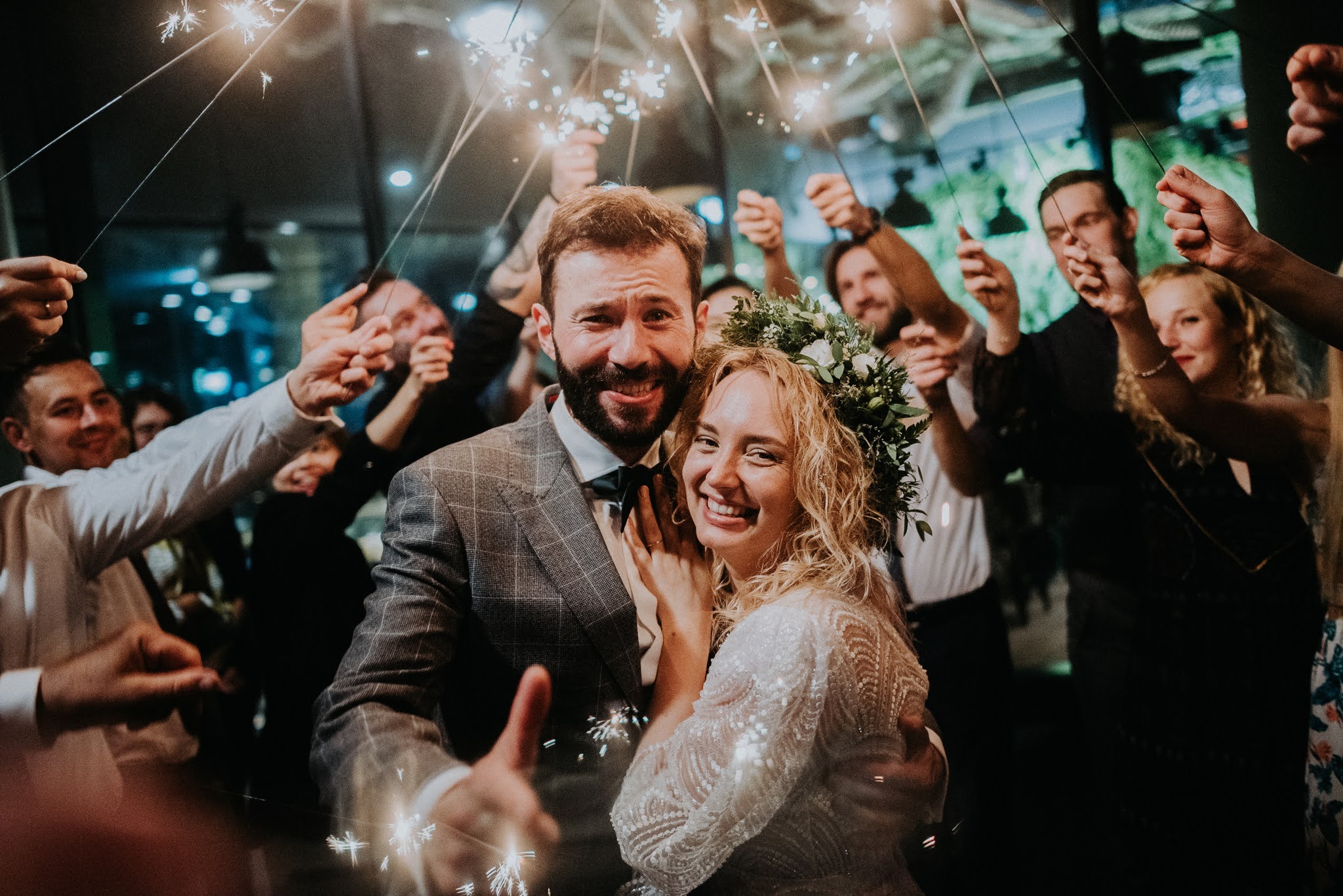 Uśmiechnięta para młoda otoczona gośćmi weselnymi z zimnymi ogniami, panna młoda w koronie z kwiatów i sukni z koronkowymi rękawami, pan młody w kraciastej marynarce i muszce.