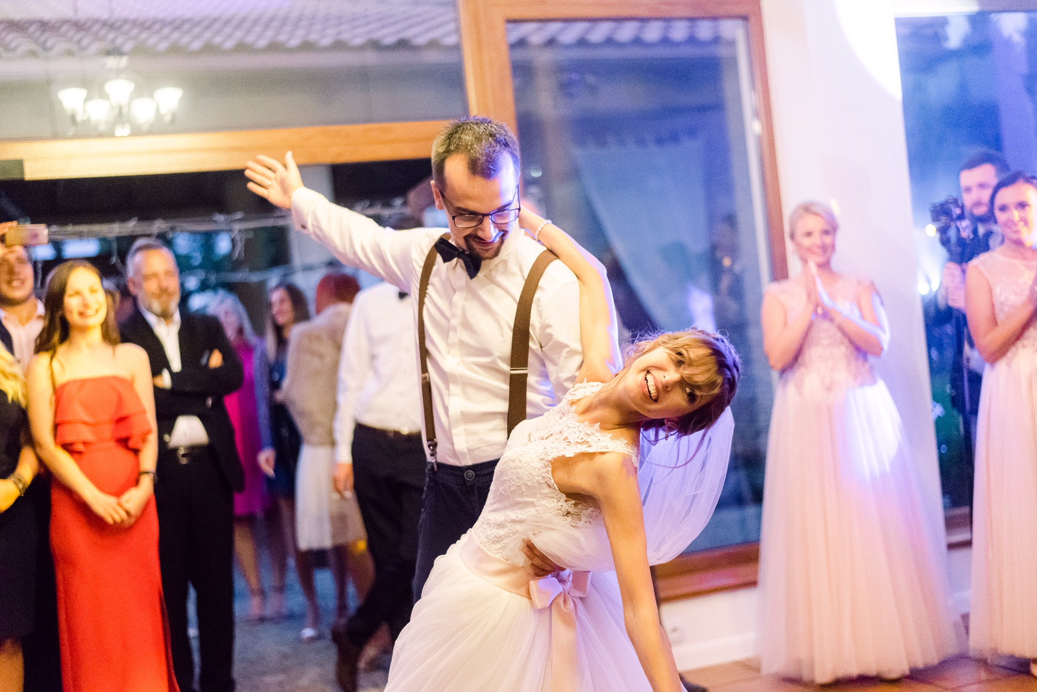 Para młoda w tańcu podczas wesela, pan młody z muchą i szelkami, uśmiechnięta panna młoda w koronkowej sukni i welonie, goście w tle.