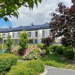 Pracownia Architektoniczna ART-PRO Sp.z.o.o - Nowoczesny budynek wielorodzinny z panelami słonecznymi na dachu, otoczony bujną zielenią, kwitnącymi krzewami i drzewami liściastymi.