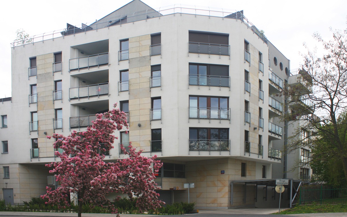 Wielopiętrowy budynek mieszkalny w Jadwisinie z balkonami i elementami elewacji wykończonymi kamieniem, z kwitnącym drzewem na pierwszym planie.