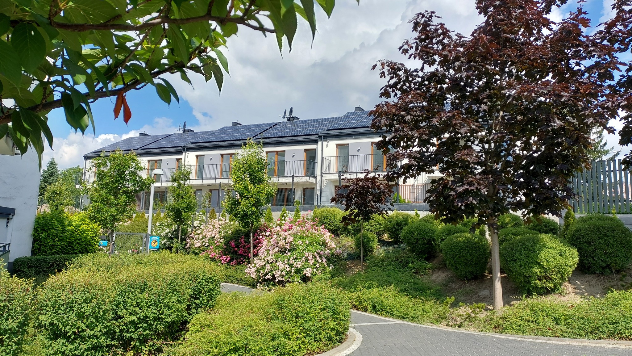 Nowoczesny budynek wielorodzinny z panelami słonecznymi na dachu, otoczony bujną zielenią, kwitnącymi krzewami i drzewami liściastymi.