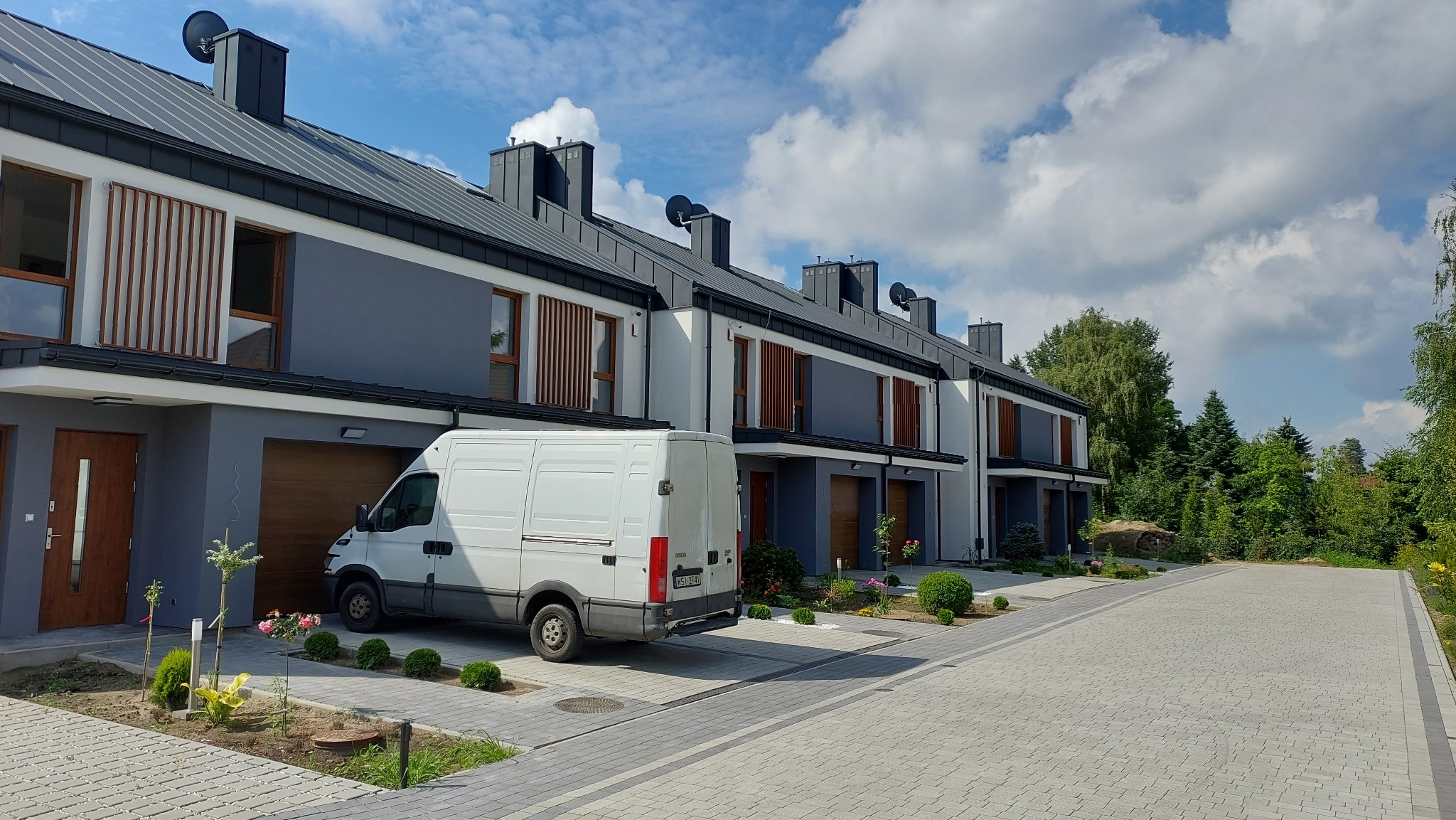 Szereg identycznych, nowoczesnych domów jednorodzinnych z szarymi elewacjami i drewnianymi akcentami, biały samochód dostawczy zaparkowany przed jednym z garaży, świeżo założony trawnik i rośliny.