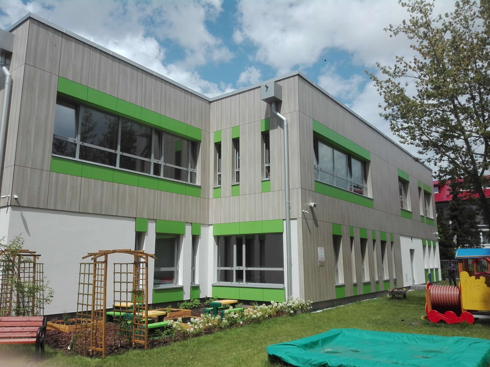 Nowoczesny budynek przedszkola z drewnianą elewacją i zielonymi obramowaniami okien, otoczony zielonym trawnikiem i elementami placu zabaw.