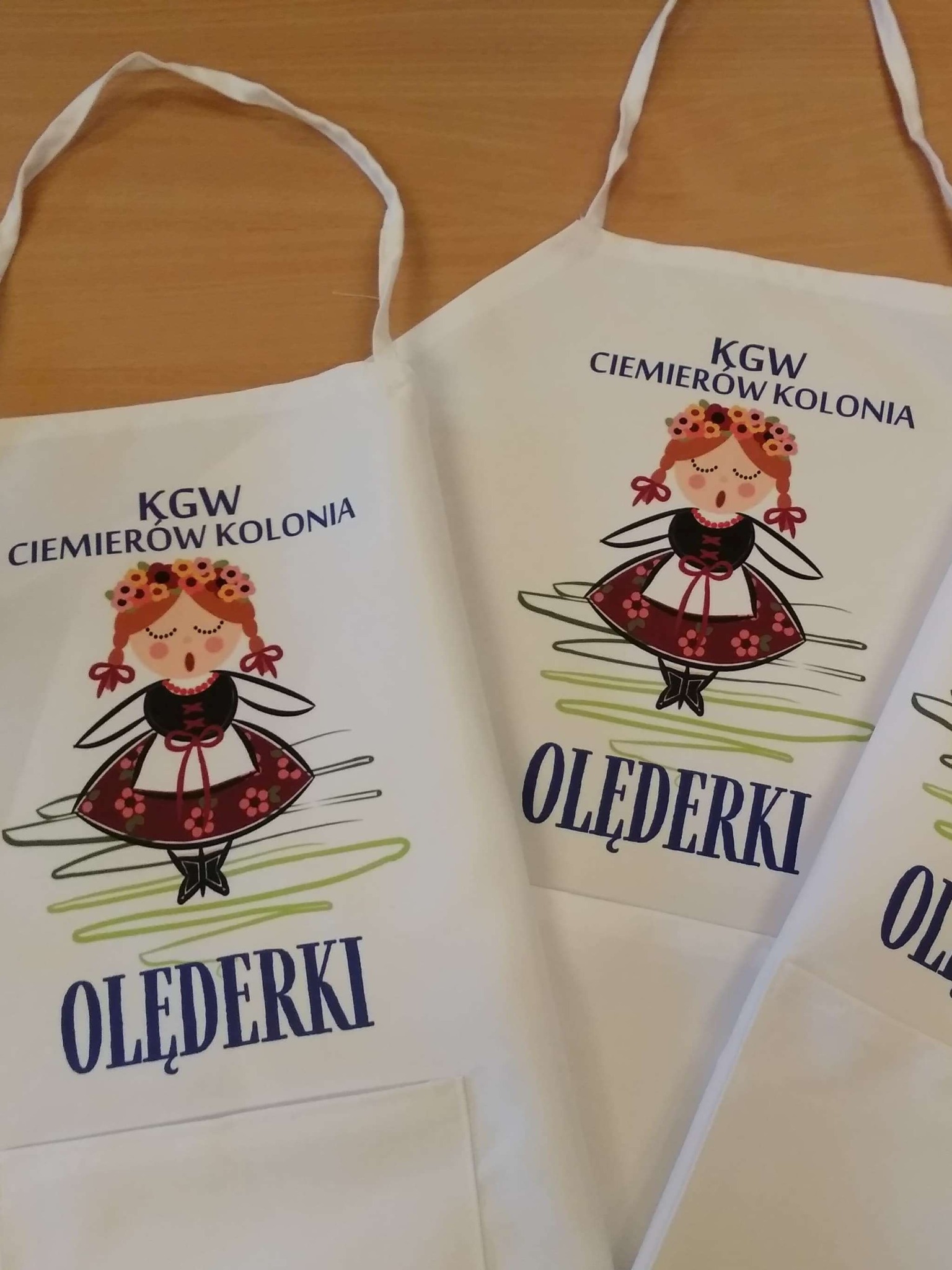 Białe fartuchy z nadrukiem ludowego motywu dziewczyny w stroju, napisu 'KGW Ciemierów Kolonia' oraz nazwy zespołu 'Olęderki'.