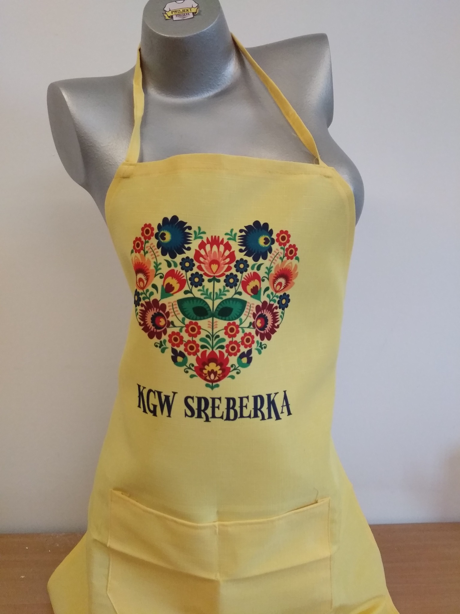 Żółty fartuch kuchenny z folkowym wzorem kwiatowym w kształcie serca i napisem KGW SREBERKA, prezentowany na manekinie.
