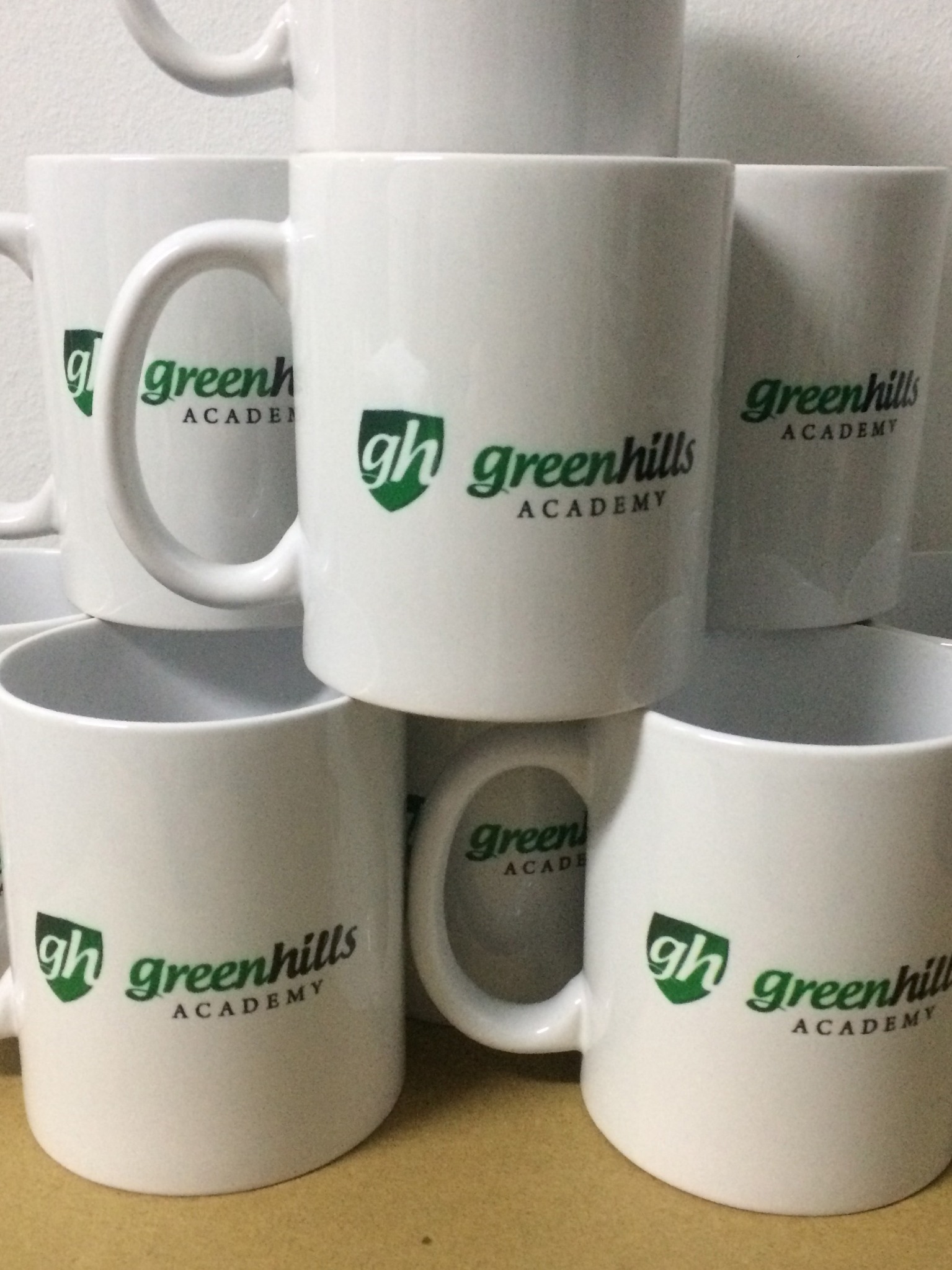 Stos białych ceramicznych kubków z zielonym nadrukiem logo Greenhills Academy.