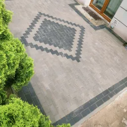 Ułożona kostka brukowa w odcieniach szarości z dekoracyjnym wzorem w kształcie rombu, otoczona niskimi, zielonymi krzewami i fragmentem elewacji budynku z oknem.