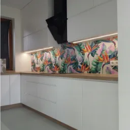 Biała, nowoczesna zabudowa kuchenna z czarnym okapem i blatem w kolorze jasnego drewna, ozdobiona panelem ściennym z motywem tropikalnych kwiatów i liści.