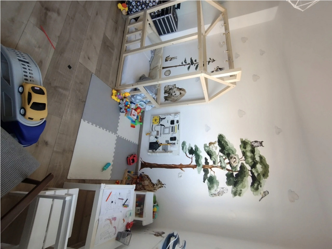 Pokój dziecięcy z łóżkiem w kształcie domku, naklejkami drzew na ścianie i interaktywną tablicą, zabawki na podłodze z puzzli piankowych.