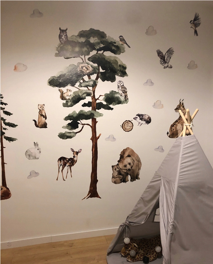 Pokój dziecięcy z naklejkami na ścianie przedstawiającymi leśne zwierzęta i drzewa, obok szary tipi z lampkami.