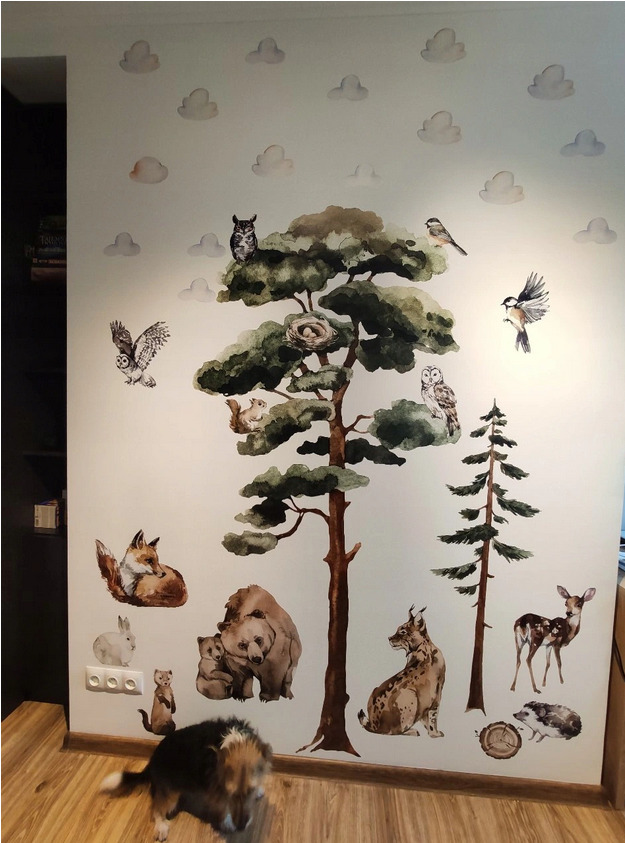 Pokój dziecięcy z naklejkami akwarelowymi przedstawiającymi leśne zwierzęta i drzewa na ścianie, stylizowane na malowane akwarelą obłoki na górze, widoczny fragment podłogi i pies w dolnej części...