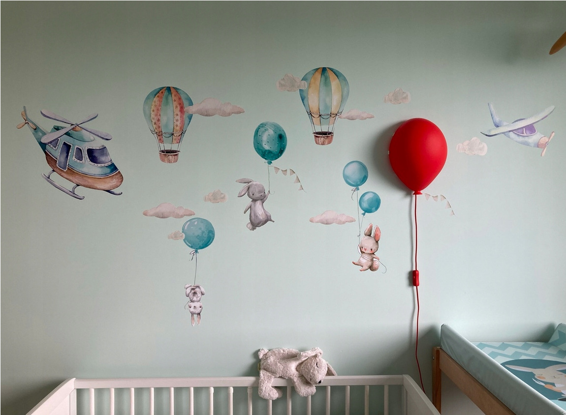 Dziecięcy pokój z pastelowymi naklejkami na ścianie przedstawiającymi helikopter, balony na ogrzane powietrze, samolot i króliki, a także lampa w kształcie czerwonego balona, białe łóżeczko...