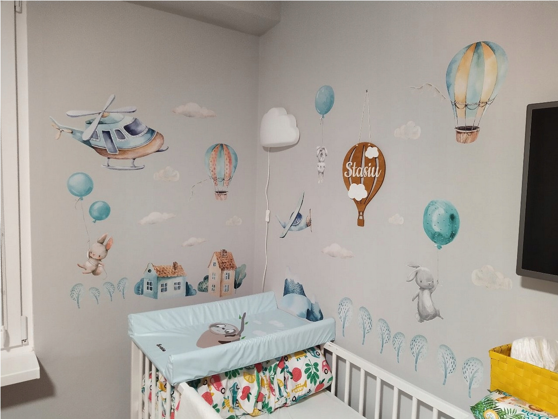 Uroczy pokój dziecięcy w Ostrowcu Świętokrzyskim z pastelowymi naklejkami na ścianach przedstawiającymi helikopter, balony, domki i zwierzątka, widoczny przewijak na łóżeczku.