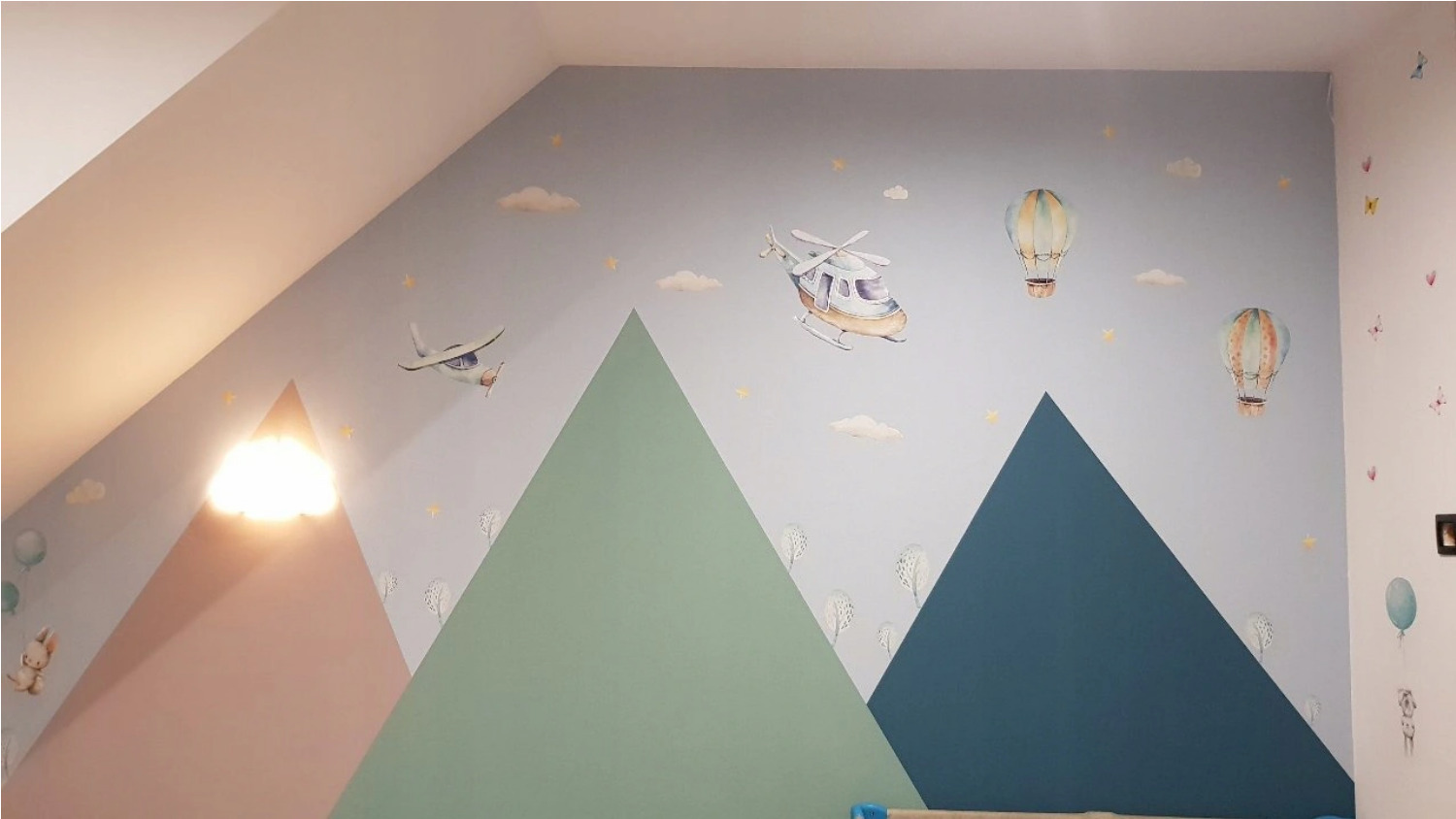 Pokój dziecięcy z pomalowaną ścianą w góry, samoloty, helikoptery i balony, dodatkowo naklejki z chmurkami i gwiazdkami, oświetlenie w kształcie chmury.