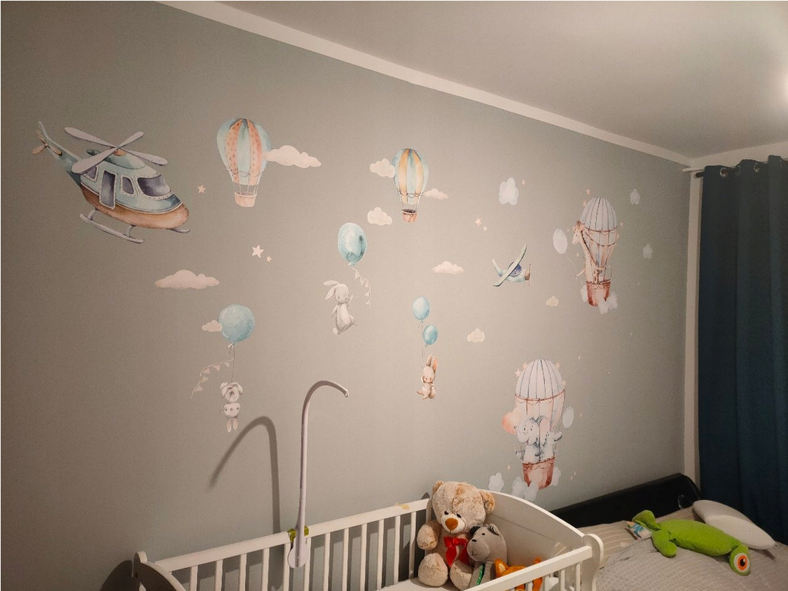 Pokój dziecięcy z szarą ścianą ozdobioną naklejkami: helikopter, balony, samolot, chmurki i gwiazdki. Białe łóżeczko z pluszowymi zabawkami.