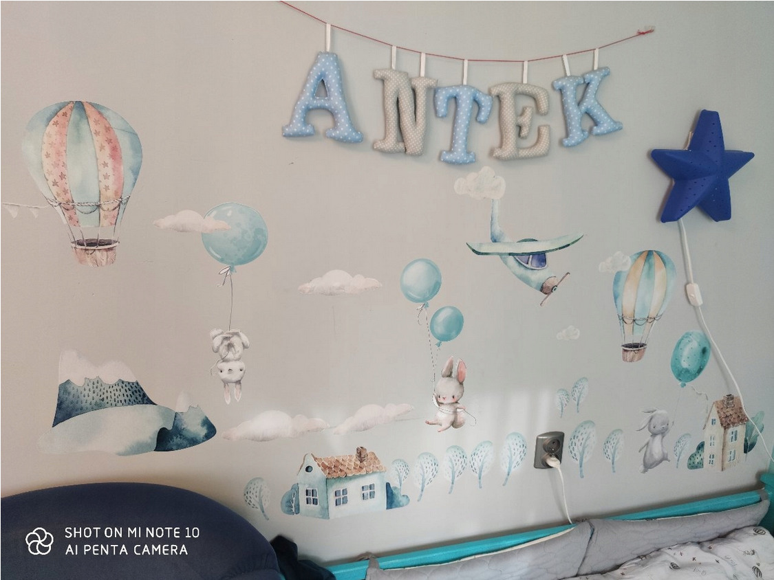 Pokój dziecięcy z pastelowymi naklejkami na ścianie: balony, samolot, zwierzęta, góry i domki. Nad łóżkiem wisi napis 'ANTEK' z materiałowych liter oraz niebieska lampka w kształcie gwiazdy.