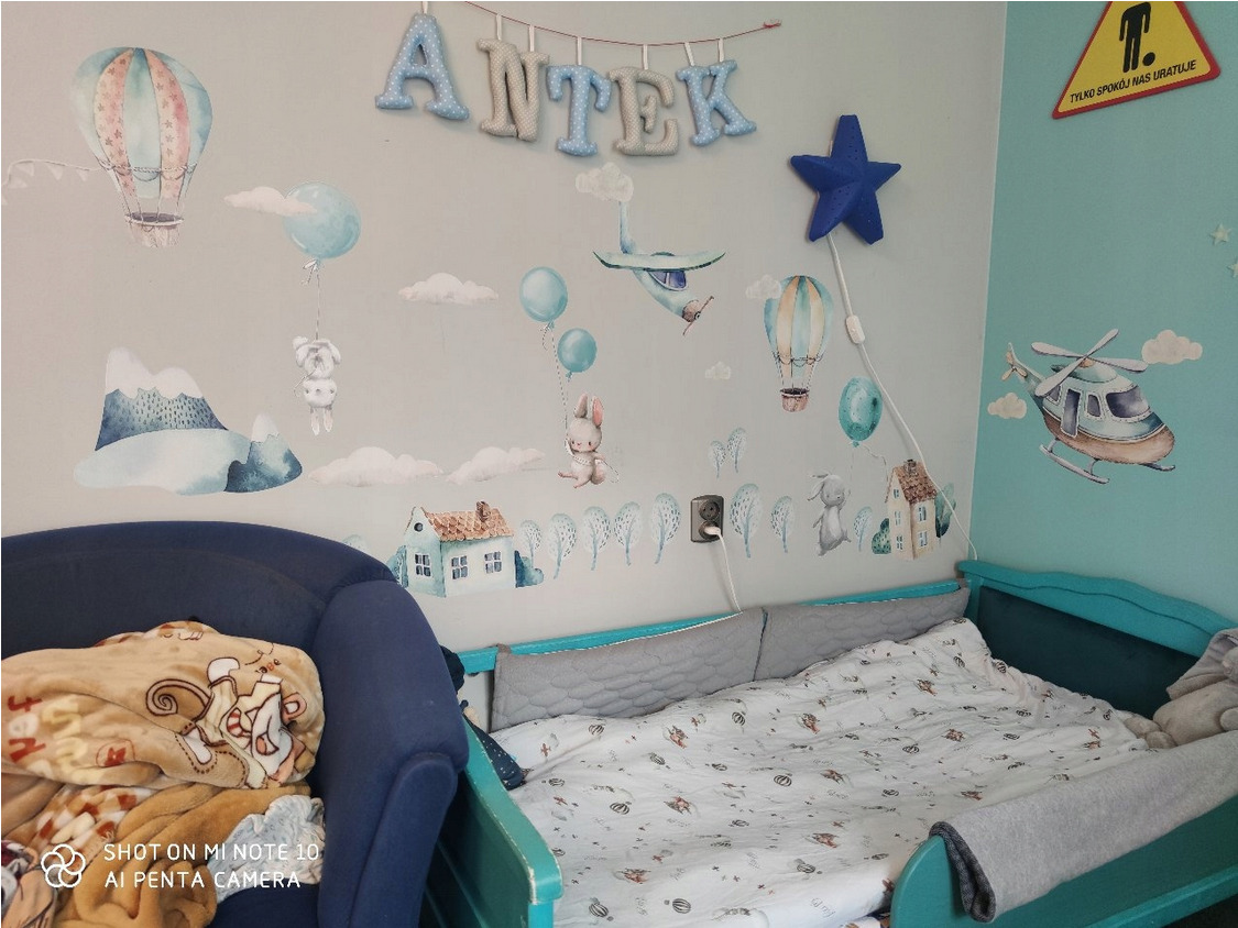 Dziecięcy pokój z turkusowym łóżeczkiem, ścianami ozdobionymi naklejkami z motywem balonów, gór, samolotów i zwierząt, oraz napisem 'ANTEK' z miękkich liter zawieszonym nad łóżkiem.