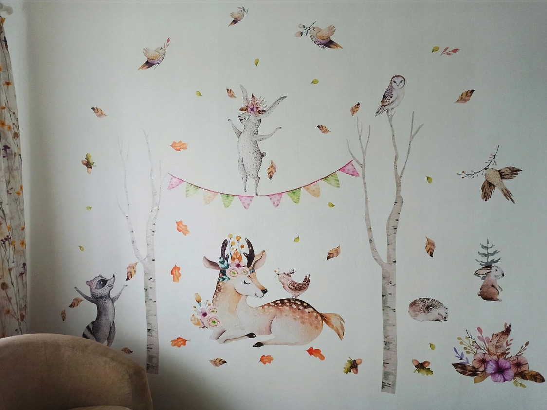 Ściana pokoju dziecięcego udekorowana naklejkami z motywem leśnych zwierząt: jelenia z kwiatami, szopa pracza, sowy, jeża i zająca na tle stylizowanych drzew i girlandy z chorągiewek.