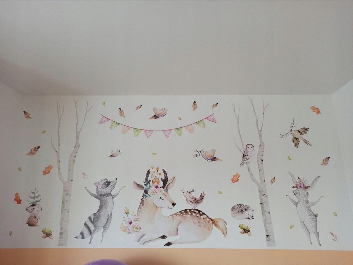 Ściana pokoju dziecięcego ozdobiona naklejkami z motywem leśnych zwierząt w stylu akwareli: jeleń z kwiatami, szop pracz, sowa na brzozie, królik i girlanda z trójkątów.
