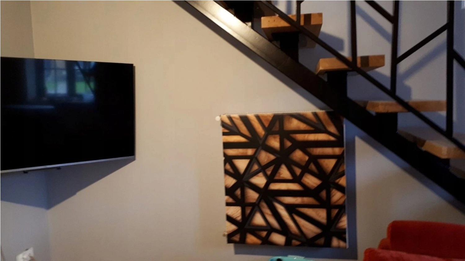 Telewizor na ścianie obok drewnianego panelu dekoracyjnego z geometrycznym wzorem, widoczne schody z metalową konstrukcją i drewnianymi stopniami.