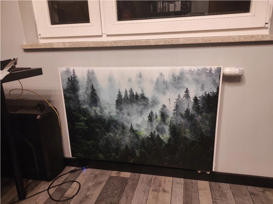 Grzejnik z nadrukiem leśnego krajobrazu, umieszczony pod oknem, widoczny fragment podłogi i biurka.