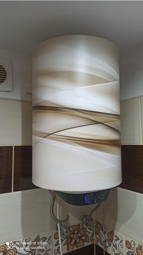 Bojler elektryczny z dekoracyjną obudową w łazience z brązowo-białymi płytkami, widoczne rury doprowadzające wodę.