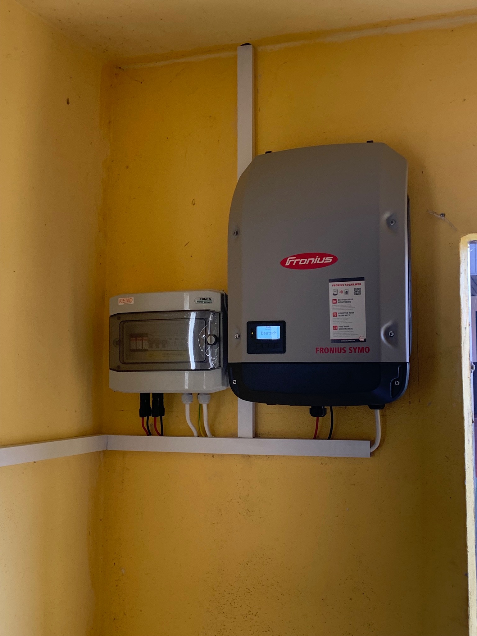 Szary inwerter Fronius Symo zamontowany na żółtej ścianie, obok skrzynka zabezpieczeń Keno, widoczne przewody i listwy maskujące.