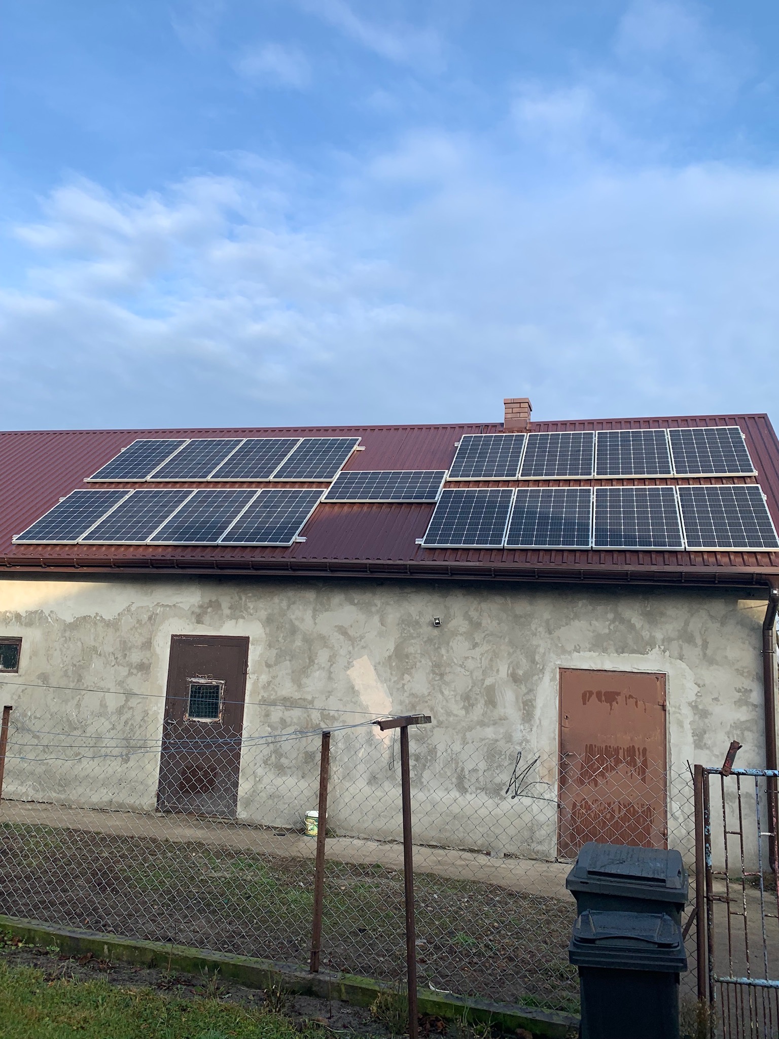 Panele fotowoltaiczne na dachu budynku gospodarczego z czerwonym dachem, widoczne dwa rzędy paneli, szary budynek z dwiema parami drzwi, przed budynkiem siatka ogrodzeniowa i czarne pojemniki...