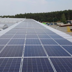 Rząd paneli słonecznych na płaskim dachu budynku przemysłowego, widok z góry, z widocznym parkingiem i drzewami w tle.