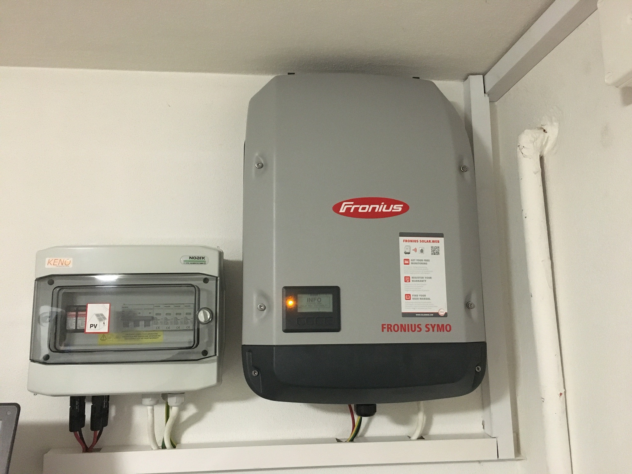 Szary inwerter Fronius Symo z włączonym pomarańczowym wskaźnikiem LED na ekranie, zamontowany obok skrzynki bezpiecznikowej KENO z naklejką PV, na białej ścianie wewnątrz budynku.