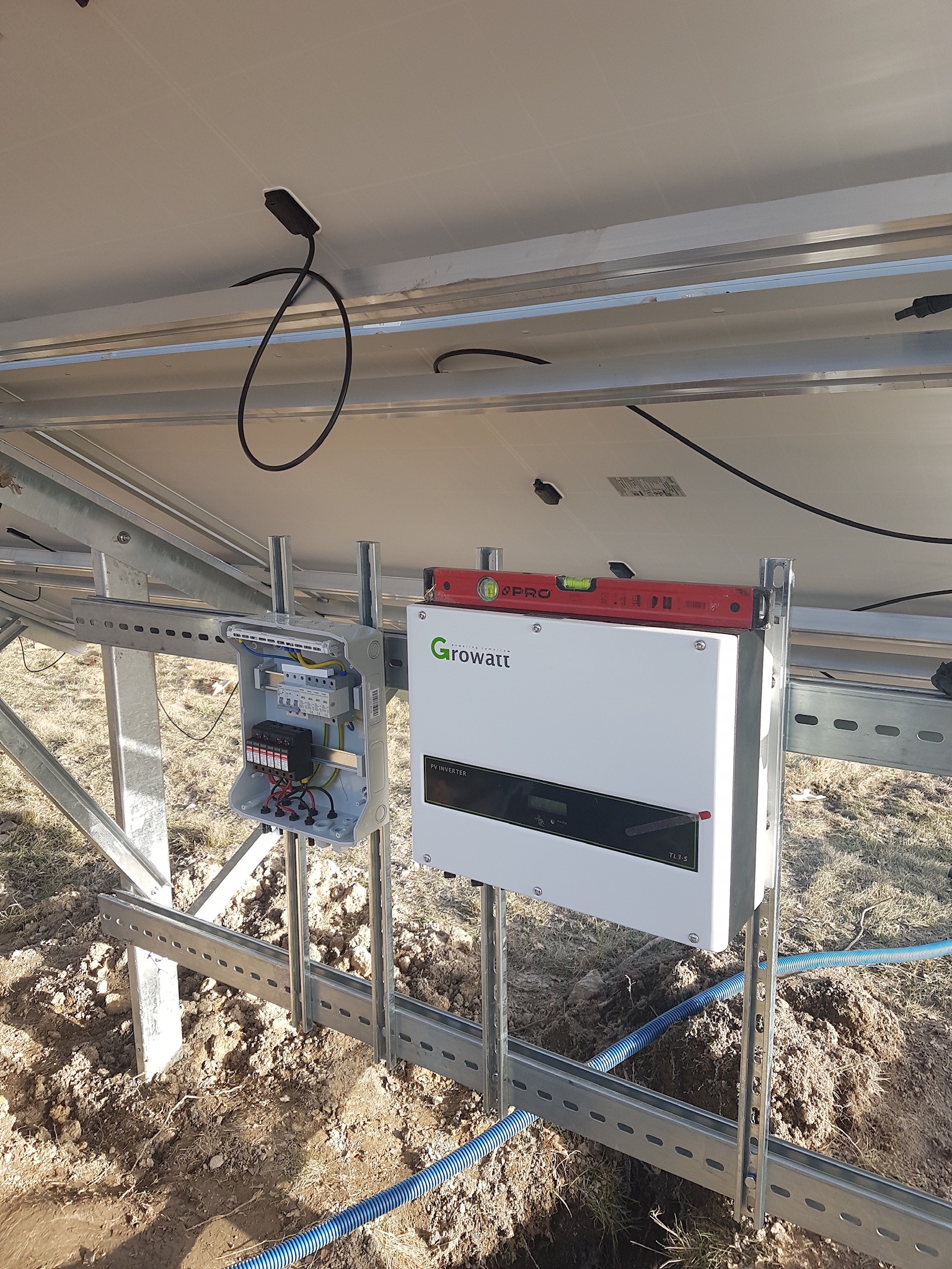 Instalacja inwertera Growatt TL3-S i skrzynki zabezpieczeń na konstrukcji wsporczej paneli słonecznych, poziomica na górze inwertera, widoczne przewody w ziemi.