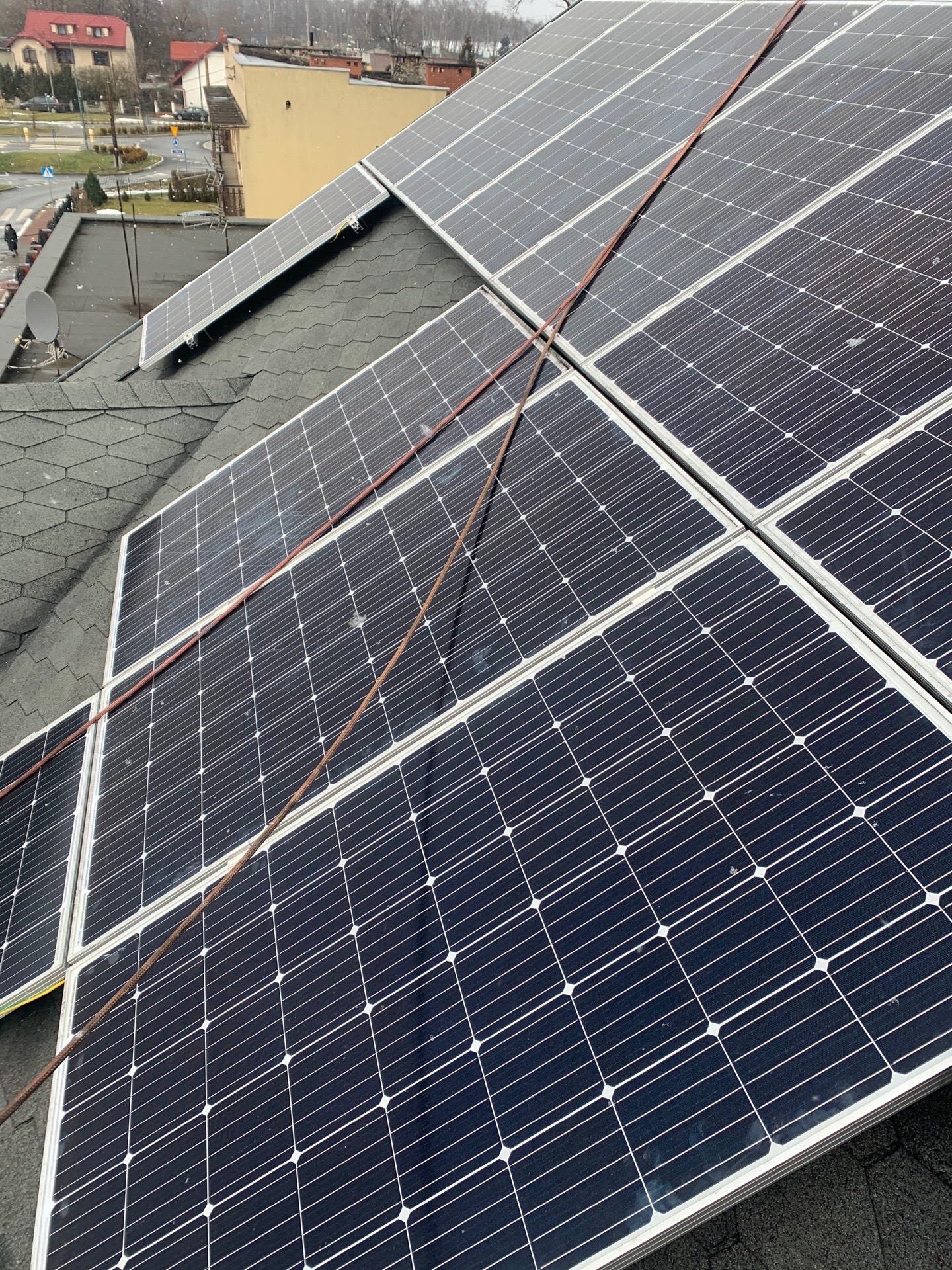 Panele słoneczne na dachu pokrytym gontem bitumicznym, zabezpieczone liną asekuracyjną; widok na zabudowę miejską w tle.