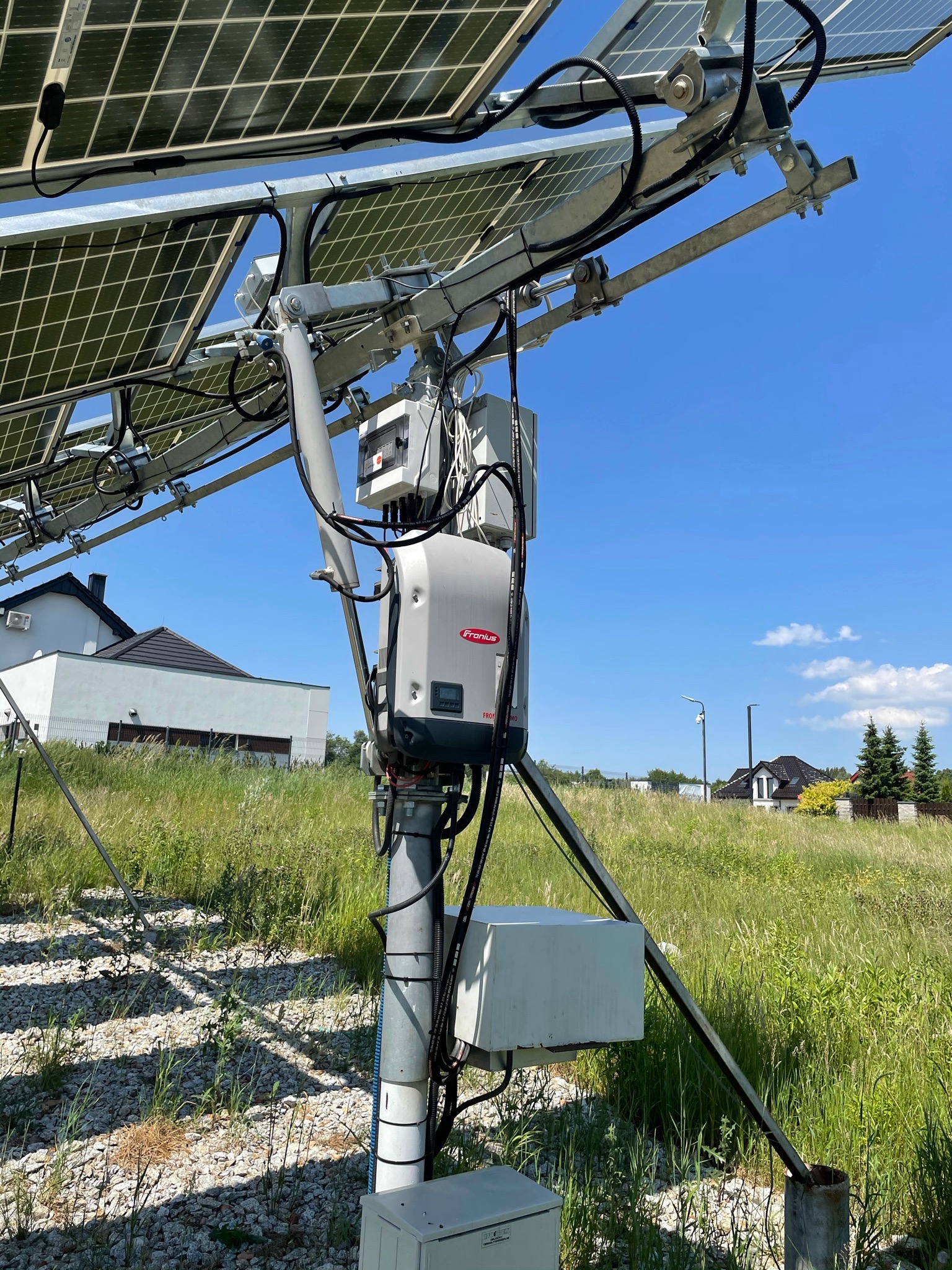 Instalacja paneli słonecznych na metalowej konstrukcji z widocznym inwerterem Fronius, zamontowana na łące z domami w tle, w słoneczny dzień.