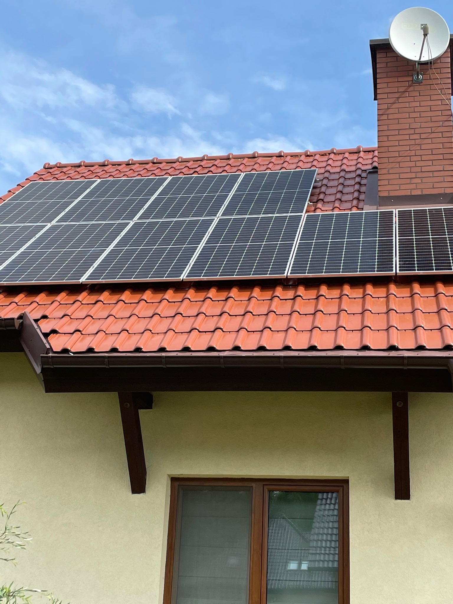Panele fotowoltaiczne na czerwonej dachówce domu jednorodzinnego z anteną satelitarną na kominie w słoneczny dzień.