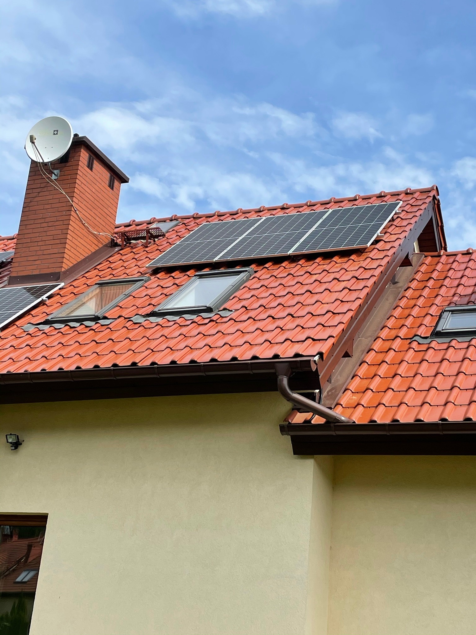 Czerwony dach z dachówki ceramicznej z zamontowanymi panelami słonecznymi i oknami dachowymi; na kominie antena satelitarna; błękitne niebo w tle.