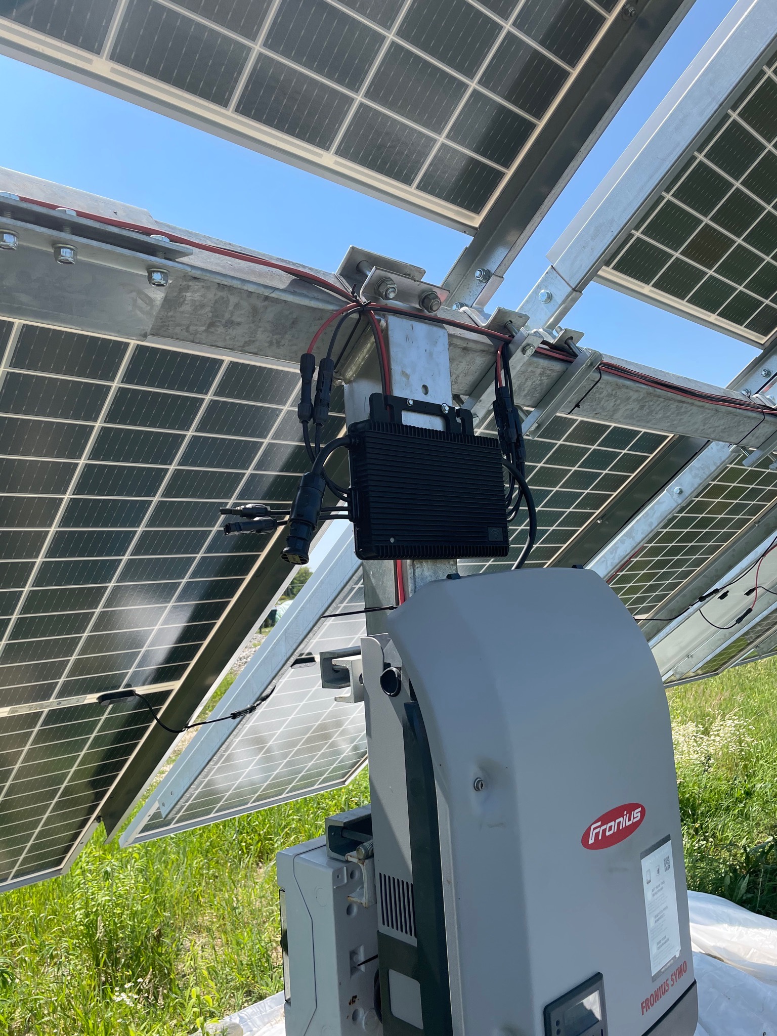 Widok od dołu na panele słoneczne i falownik Fronius Symo na metalowej konstrukcji montażowej na tle błękitnego nieba i zielonej trawy.