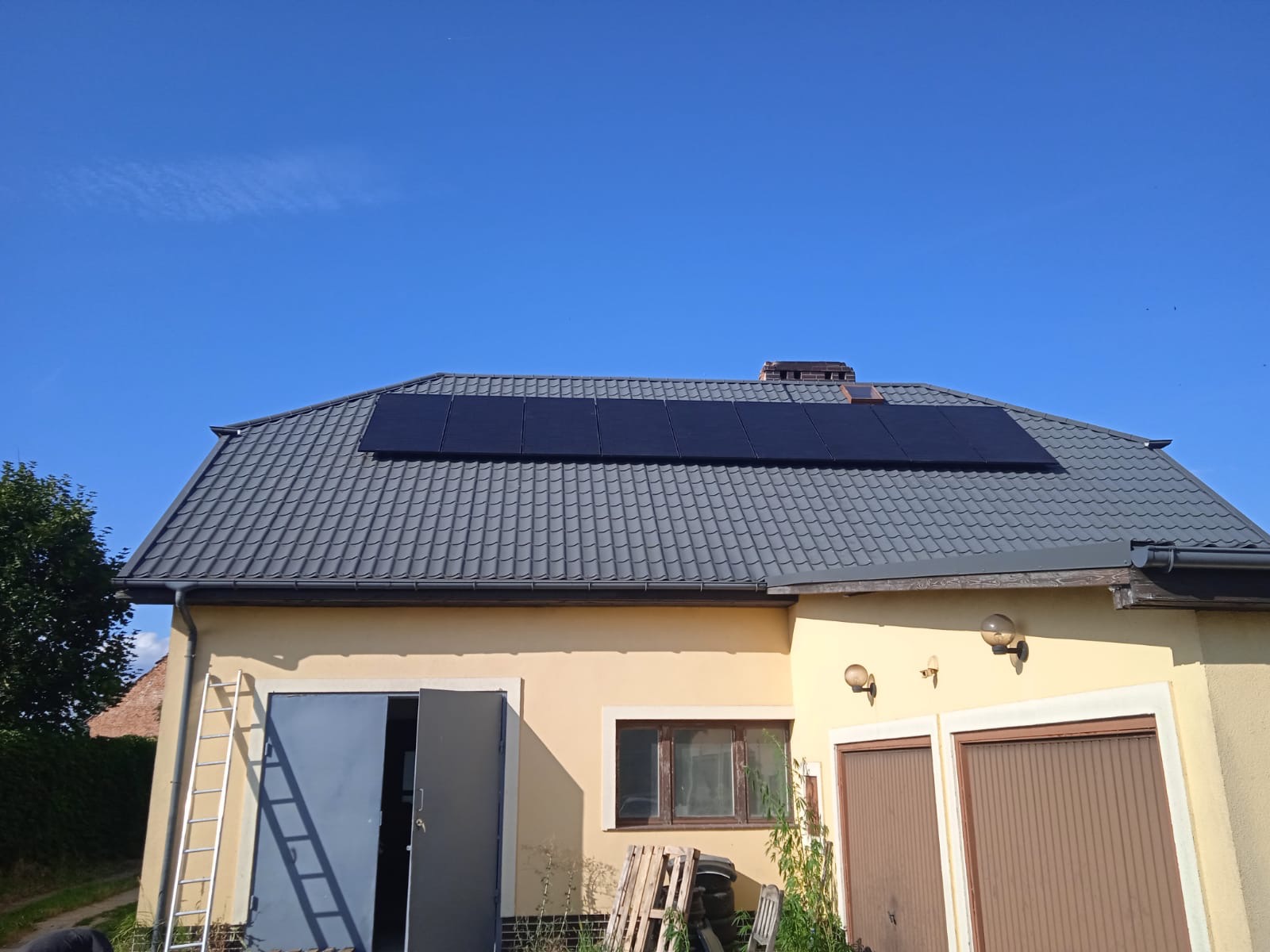 Żółty dom z szarą dachówką, na której zamontowane są czarne panele słoneczne; widoczna drabina oparta o ścianę, otwarta brama garażowa i błękitne niebo.