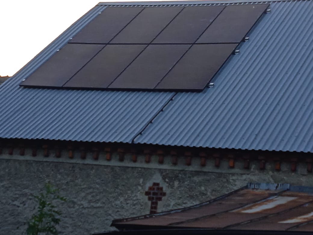 Sześć paneli słonecznych zamontowanych na falistej, metalowej blasze dachu budynku z szarą, teksturowaną ścianą poniżej. Dach sąsiedniego budynku widoczny w dolnej części kadru.