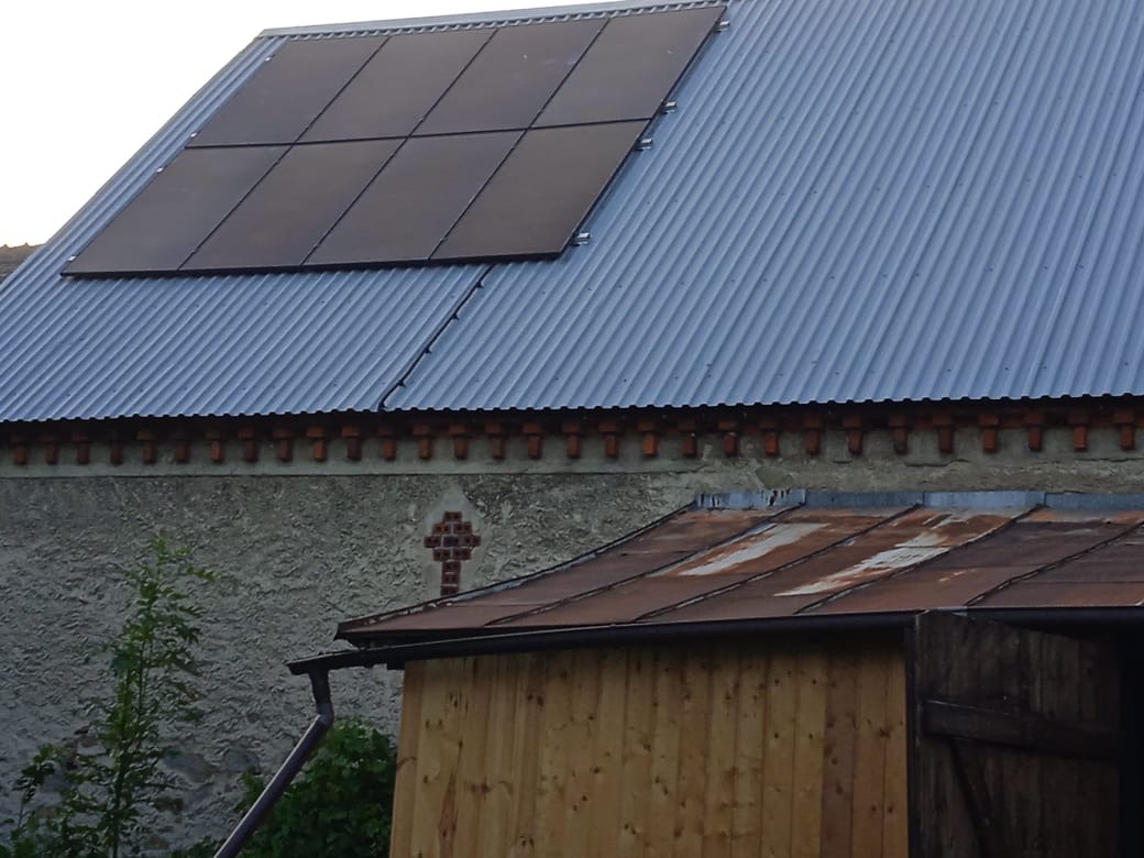 Ciemne panele słoneczne na falistym, metalowym dachu budynku gospodarczego, z widocznym fragmentem starej, otynkowanej ściany z ceglanym krzyżem i drewnianą szopą w tle.