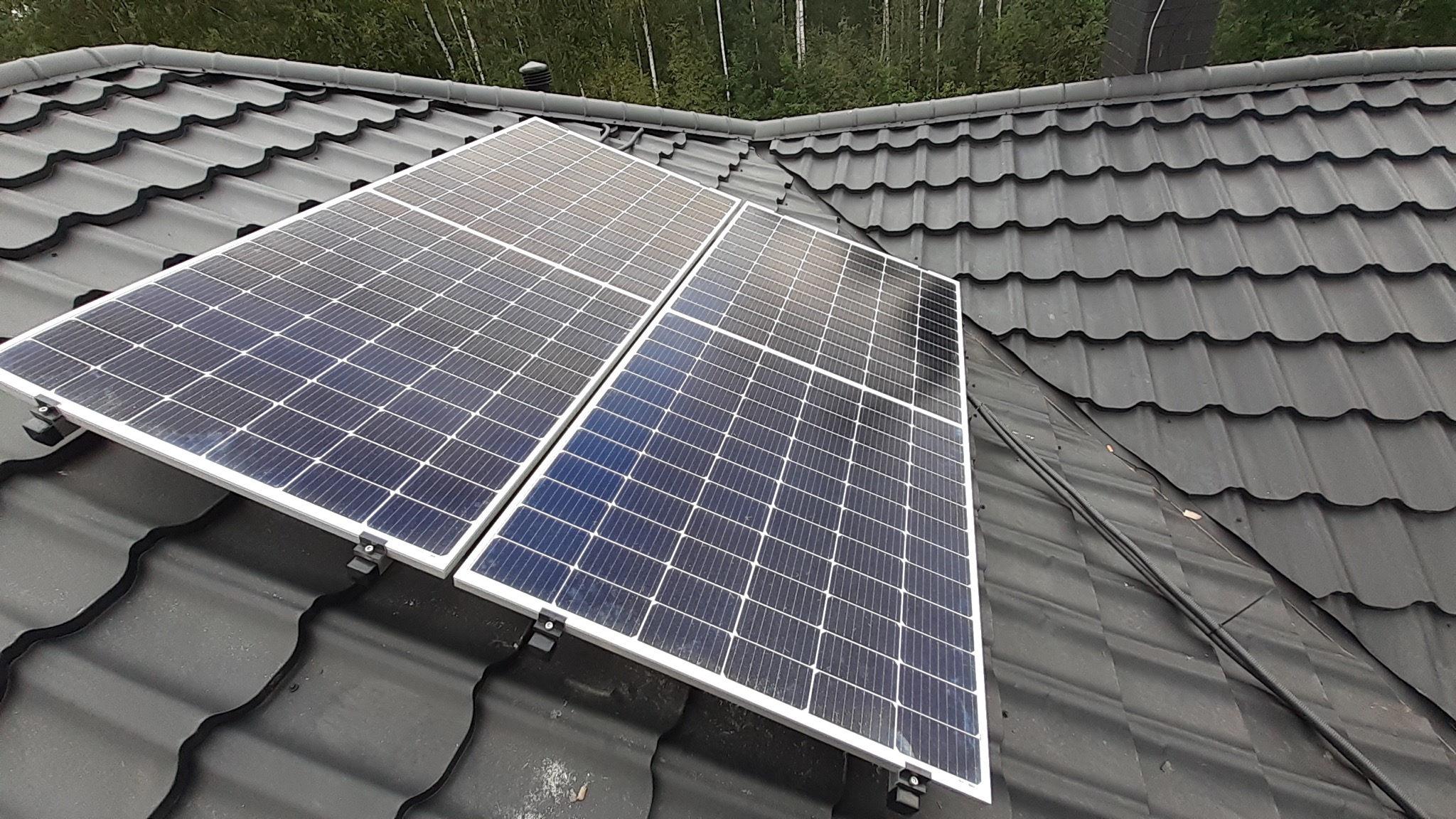 Dwa panele słoneczne zamontowane na ciemnoszarym dachu z dachówki, widoczne metalowe mocowania i przewody.