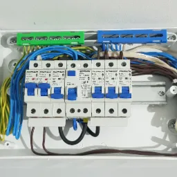 Rozdzielnica elektryczna z zainstalowanymi bezpiecznikami Schrack, widoczne okablowanie w kolorach niebieskim, żółtym, zielonym i brązowym, zamocowane na szynach DIN w białej obudowie.